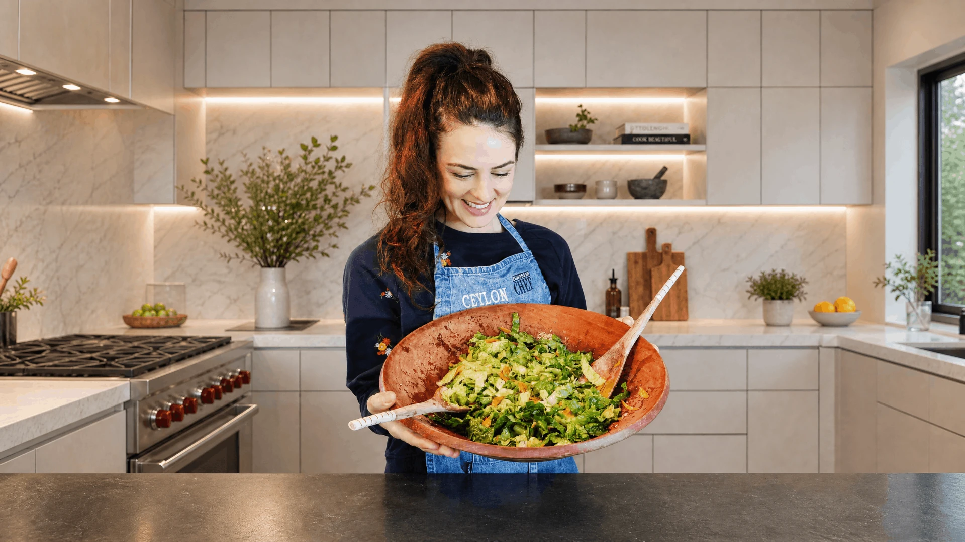 Ceylon Rose smiling in a denim apron, holding out a large terracotta bowl of chopped green salad
